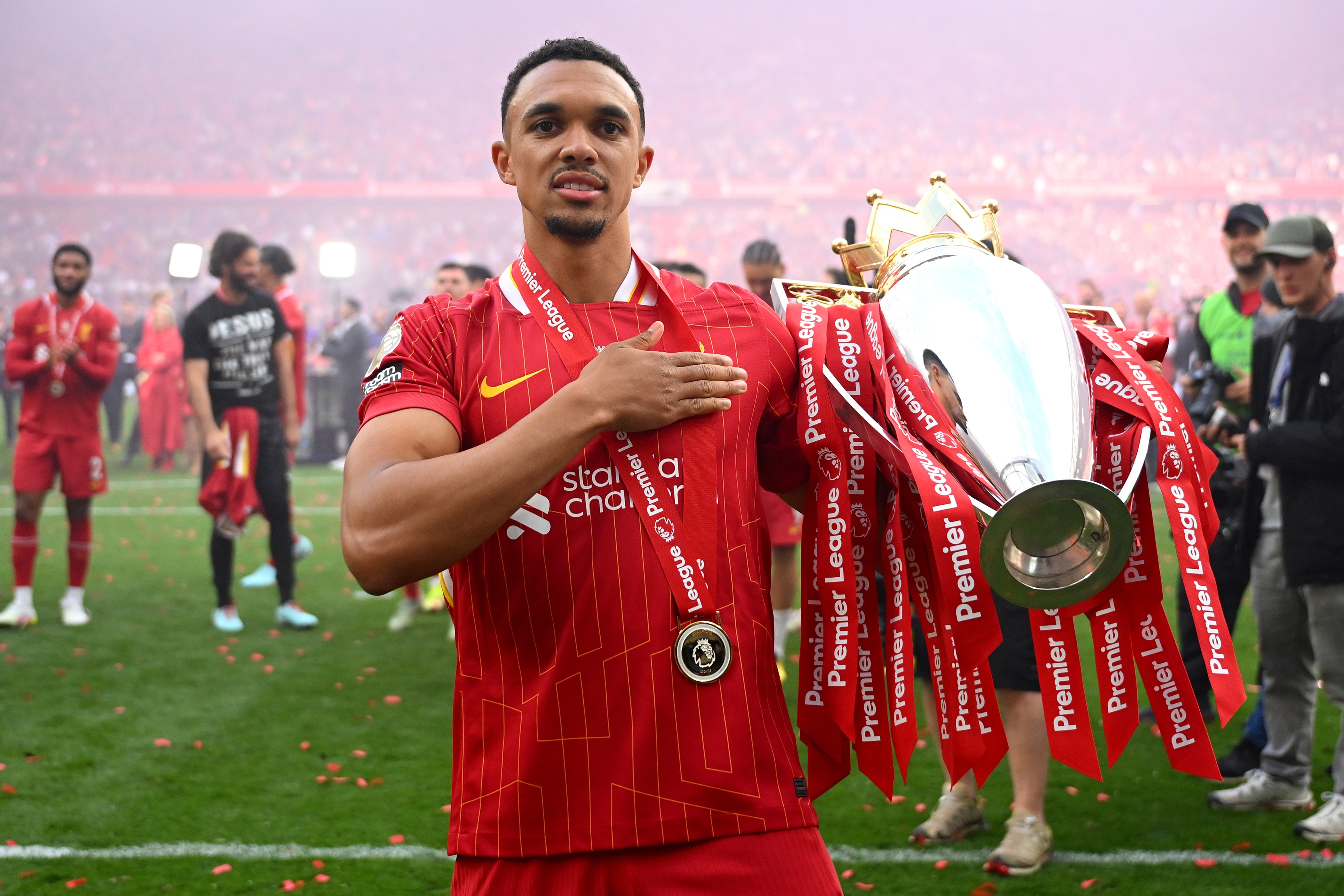 Real Madrid defender Trent Alexander-Arnold wears a red Liverpool shirt and pats the liverbird badge while holding the Premier League trophy.